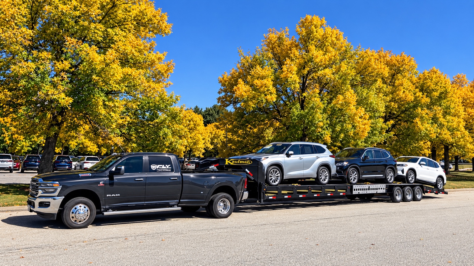 Fly Transport RAM pickup and open Kaufman trailer carrying three vehicles beneath autumn trees